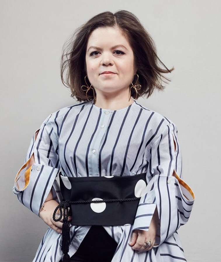 Person with shoulder-length brown hair wearing a striped shirt and a large black belt stands against a plain background.