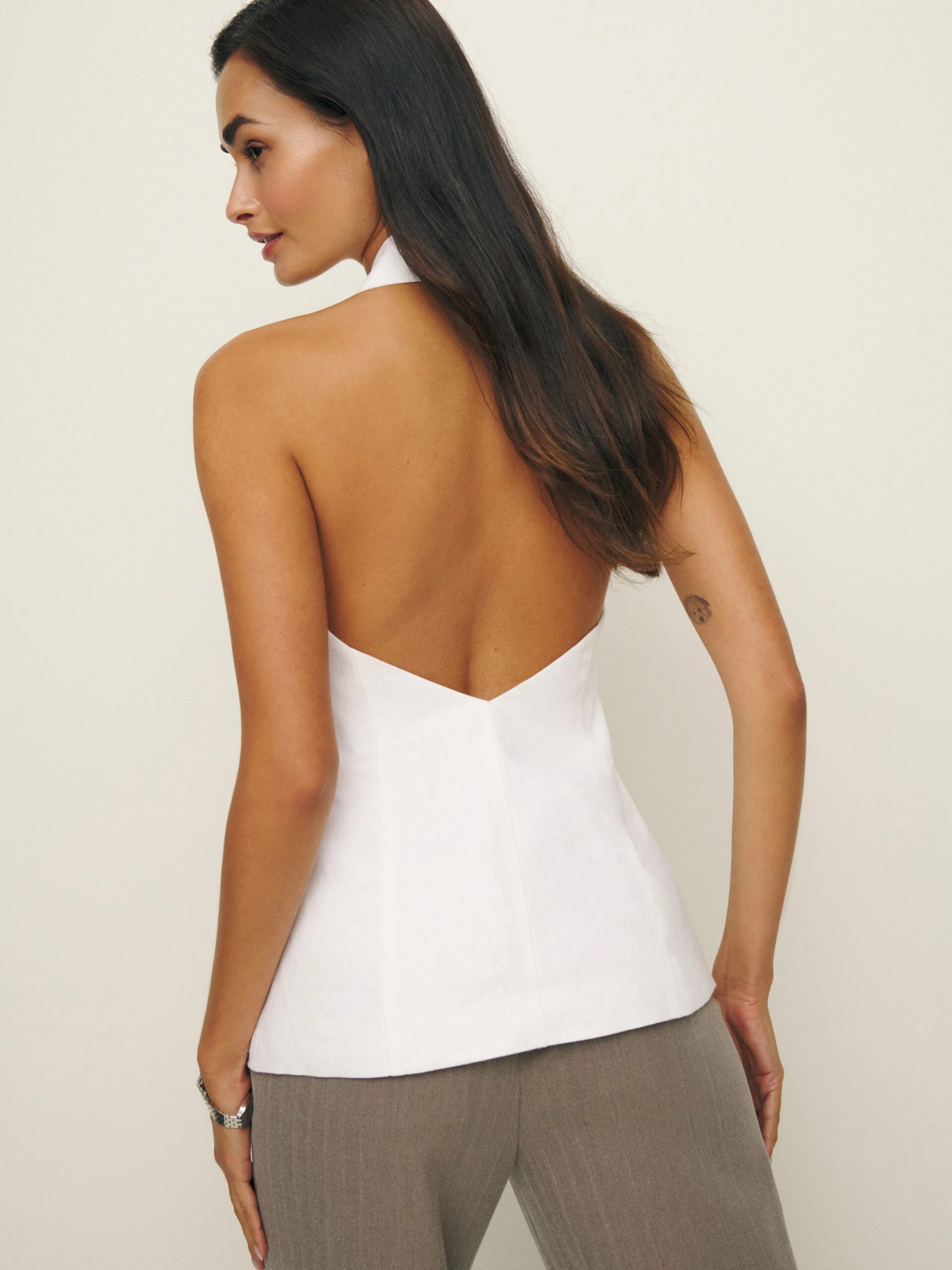 A woman with long hair is shown from the side and back wearing a sleeveless, backless white top and gray pants.