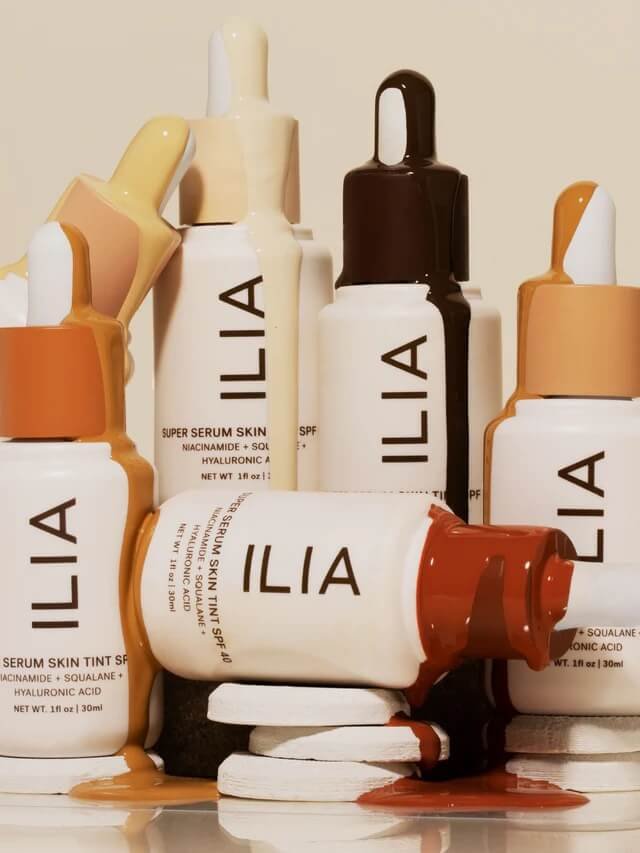 Bottles of ILIA Super Serum Skin Tint with different shades of liquid foundation spilling over white stones.