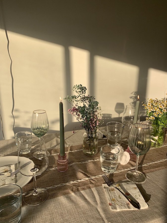 Table setting with plates, glasses, cutlery, and two lit candles on a beige tablecloth. Sunlight creates shadows on the wall.