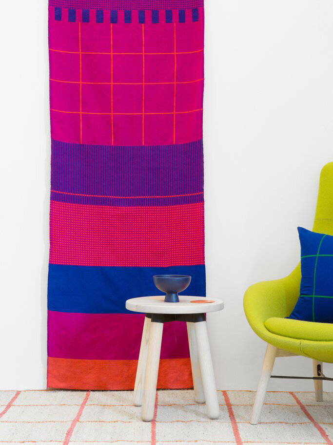 The Harar Wall Hanging in bright fuchsia, orange and blue, displayed above a side table and chair. 