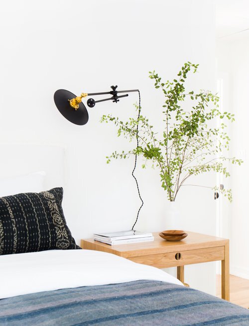A minimalist bedroom scene features an organic mattress on the bed, complemented by a small wooden side table, a wall-mounted lamp, and a leafy plant in a vase.