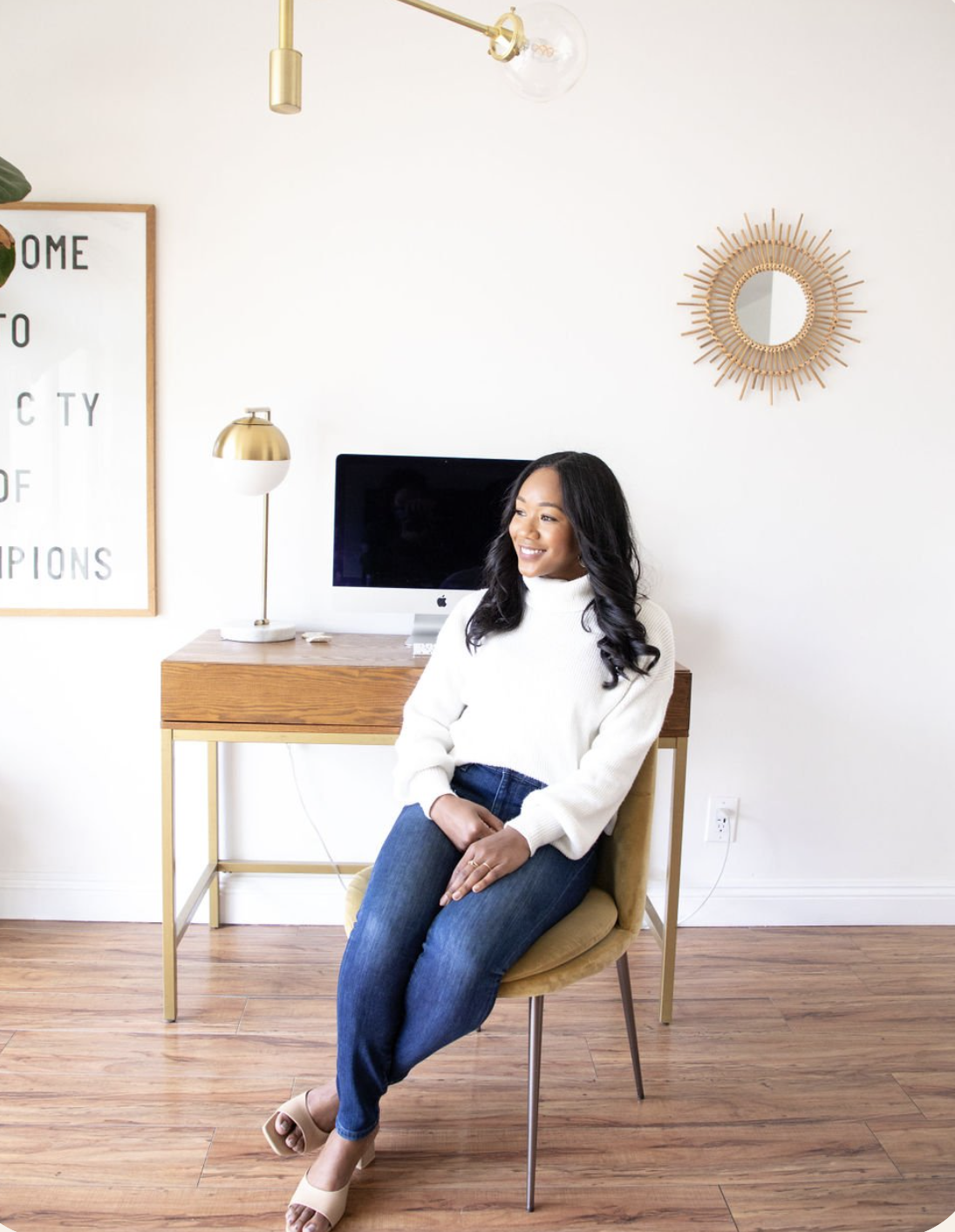 A woman in a white sweater and jeans sits on a chair in a home office with a wooden desk, computer, and wall decor.