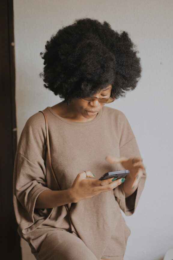 A person with curly hair and glasses is wearing a loose, beige outfit and using a smartphone while standing indoors against a plain, light-colored wall.