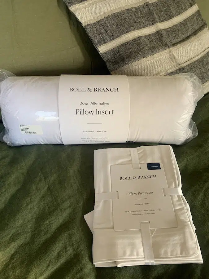 A Boll & Branch down alternative pillow insert and organic pillow protector in their packaging on a bed.