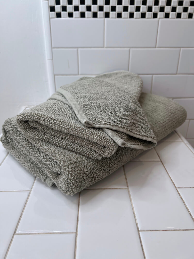 A neatly folded gray organic towel sits on a white tiled bathroom countertop with white subway tiles and a black-and-white mosaic accent in the background.