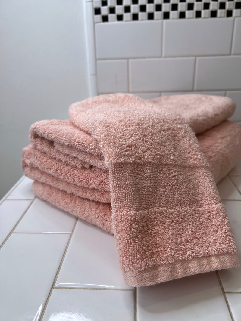 A stack of folded organic pink towels placed on a white tiled surface in a bathroom with white tiled walls.