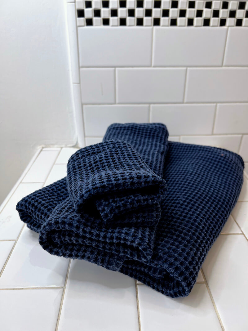 Two folded dark blue organic towels with a waffle-weave texture are stacked on a white tiled bathroom surface, with a white tiled wall in the background.
