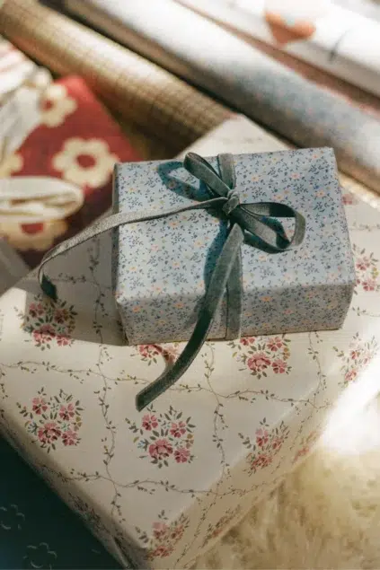 A small gift box with a blue floral pattern and gray ribbon sits on top of a larger box with a pink floral design, surrounded by rolls of wrapping paper.