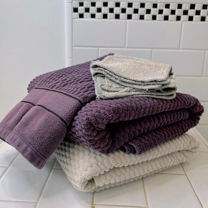 A stack of folded organic towels and washcloths in shades of purple and gray sits on a white tiled bathroom surface with a black-and-white border on the wall.
