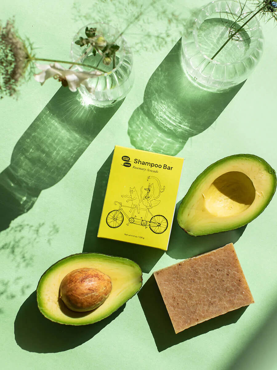 An aerial shot of a bar of Meow Meow Tweet shampoo next to its packaging, avocados, and vases with flowers. 
