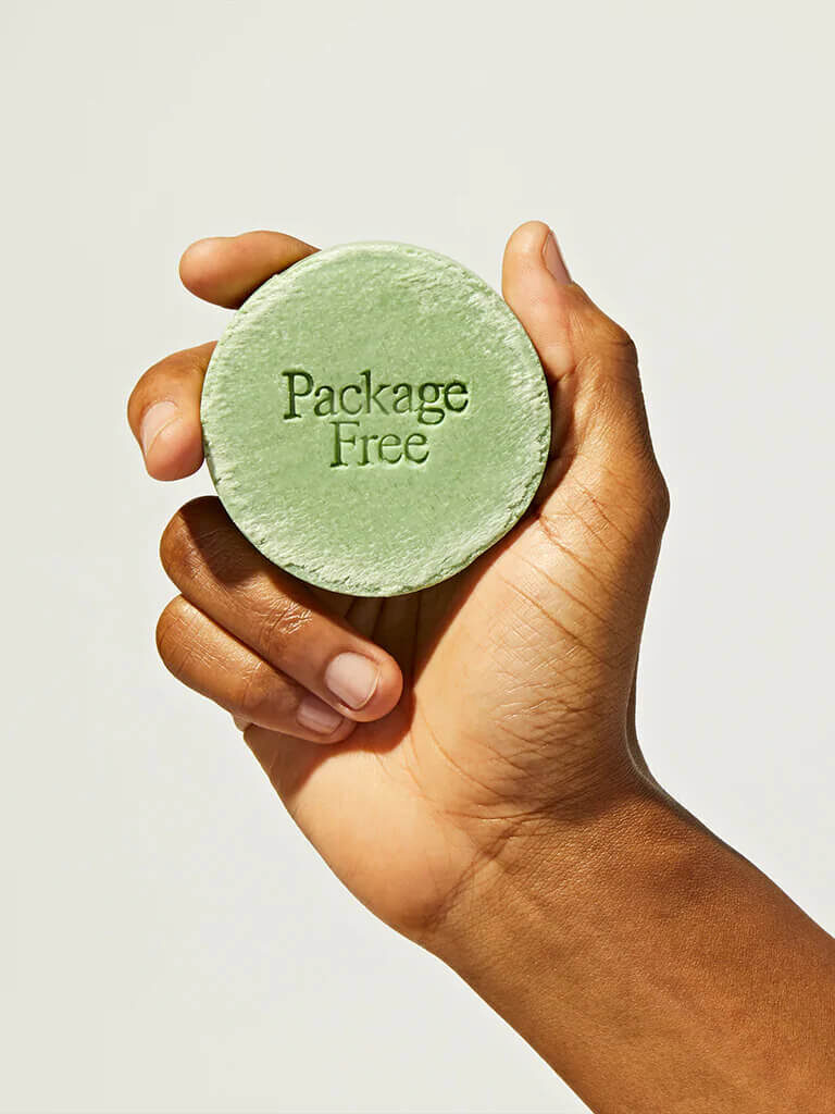A hand holding up a Package Free shampoo bar that is green and circular. 