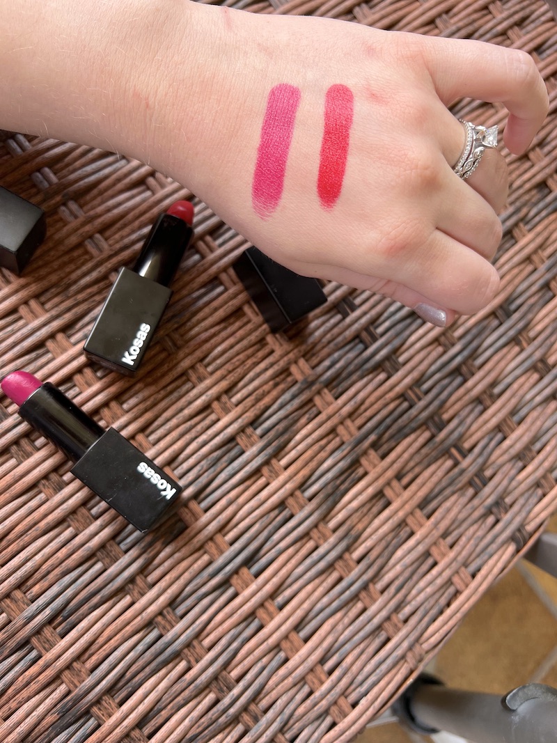 A hand displays two red lipstick swatches on the wrist. Three open Kosas lipsticks lie on a wicker surface below the hand.
