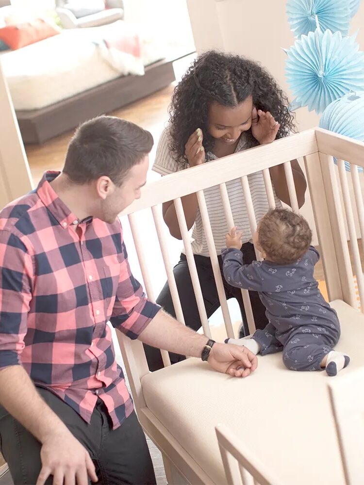 A woman and man play peek-a-boo with a baby in a Naturepedic crib mattress.