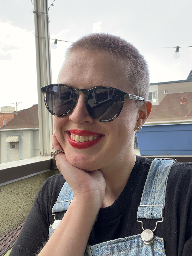 Person with short hair, wearing sunglasses, a black shirt, and denim overalls, smiling with hand resting on cheek, seated outdoors with buildings and string lights in the background.