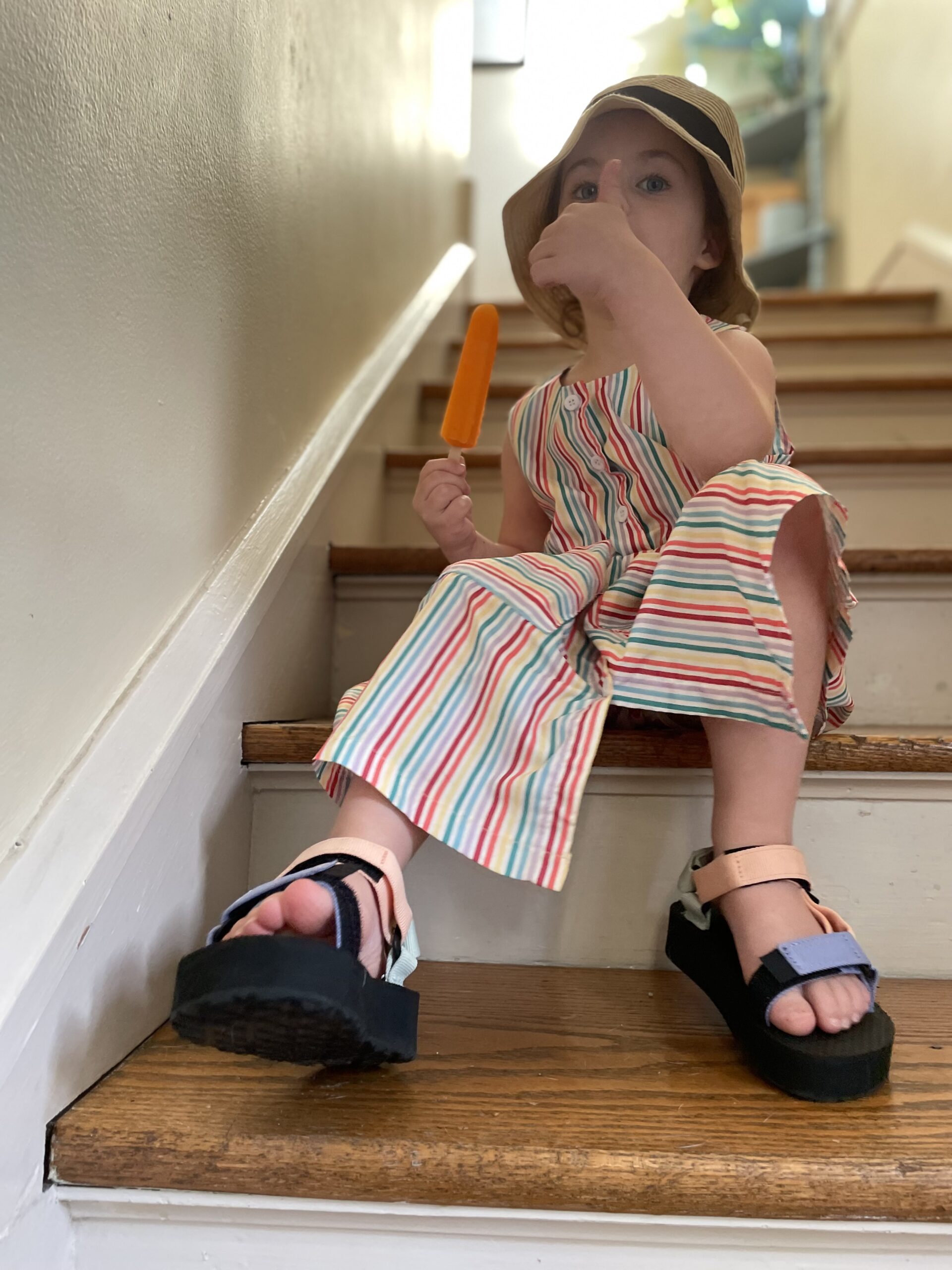A little girl in a dress, Tevas, and a bucket hat makes a thumbs up.