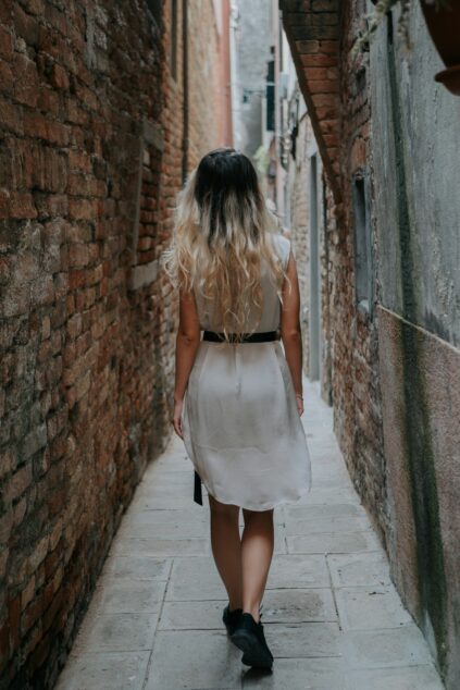 A woman with long, wavy blonde hair in a white dress walks down a narrow, brick alleyway.