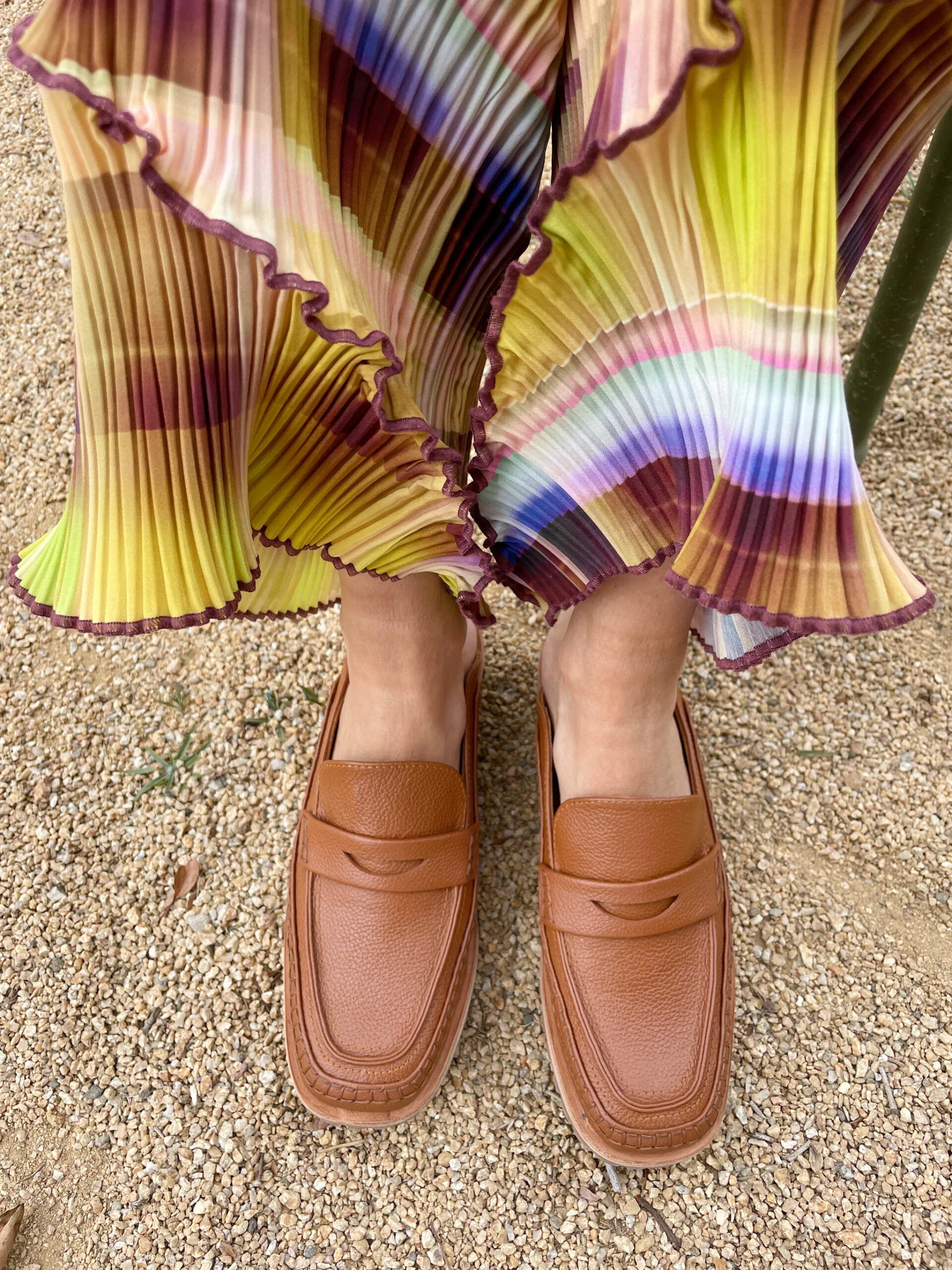 A pair of brown penny loafer-style mules on a model's feet, a multicolored pleated skirt fanning above it.