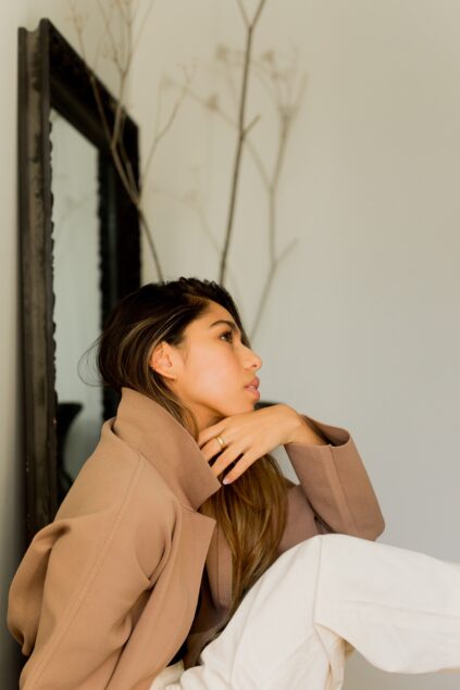 Woman in a brown coat and white pants sits by a mirror, looking to the side, with a bare branch in the background.