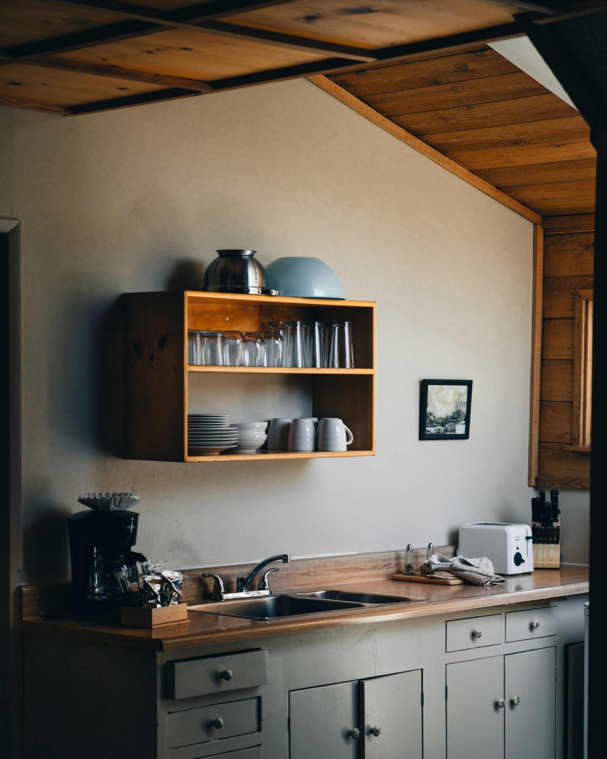 A small kitchen features a sink, countertop appliances, a wall-mounted wooden shelf with dishes and glasses, and neutral colors with wood accents.
