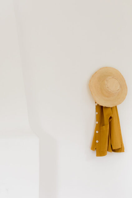 A straw hat and a mustard yellow button-up shirt hang on a white wall.