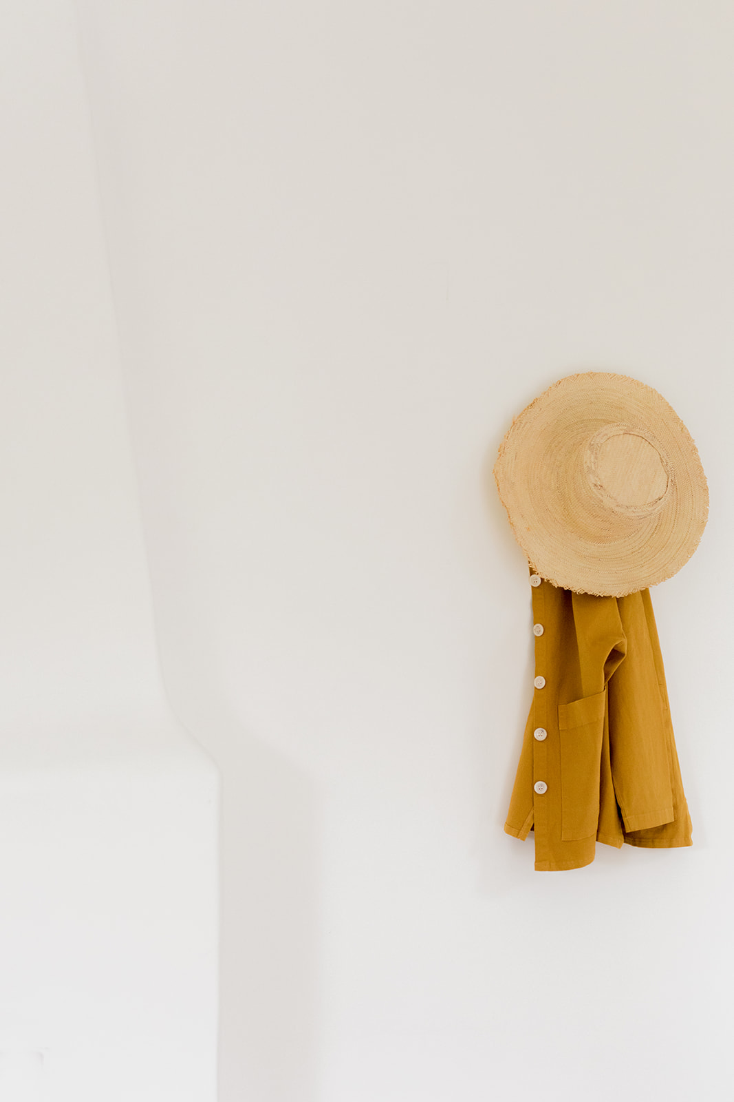 A straw hat and a mustard yellow button-up shirt hang on a white wall.