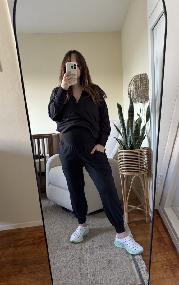 Person taking a mirror selfie in a room, wearing a black outfit and white clogs, with a plant and armchair visible in the background.