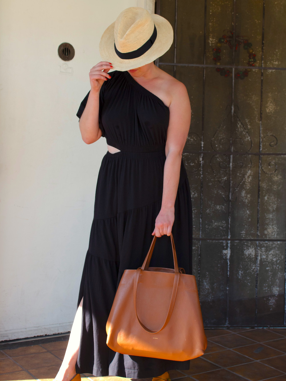 Person wearing a black one-shoulder dress and straw hat stands in front of a metal door, holding a large brown tote bag.