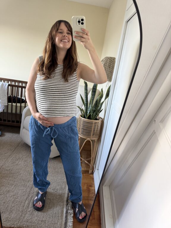 A woman stands in a bedroom taking a mirror selfie, smiling, with one hand on her belly. She is wearing a striped top, blue pants, and sandals. A crib and plant are in the background.
