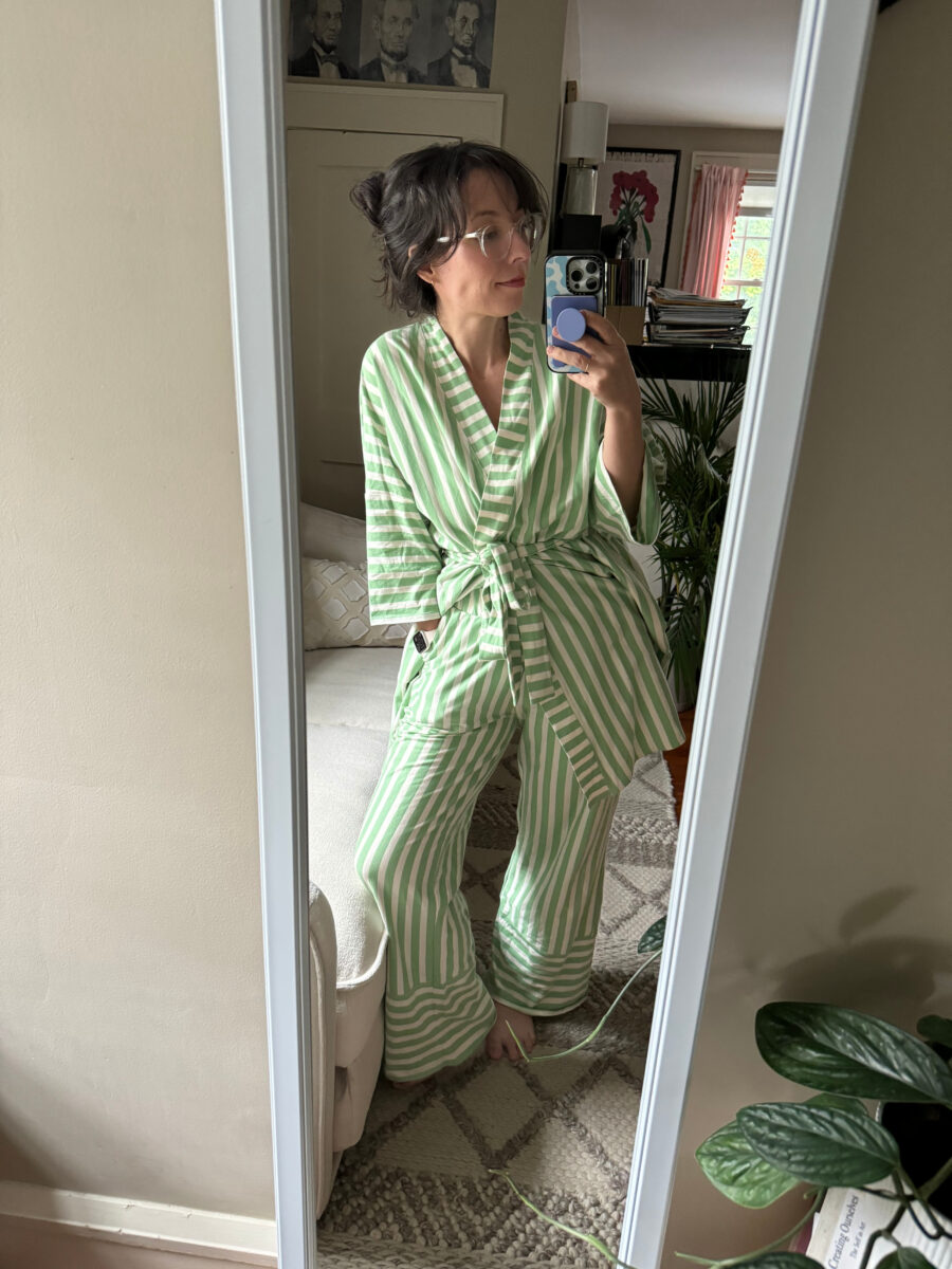Person wearing green and white striped pajamas stands in front of a mirror, taking a selfie in a room with framed art and plants visible in the background.
