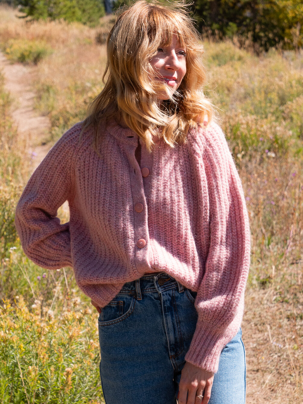 A person stands outdoors on a grassy path, wearing a pink knit sweater and blue jeans, with sunlight highlighting their hair.