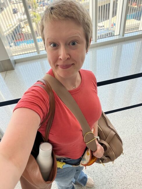 Person with short hair and a red shirt holding a brown bag and a water bottle, standing indoors near large windows and looking at the camera.