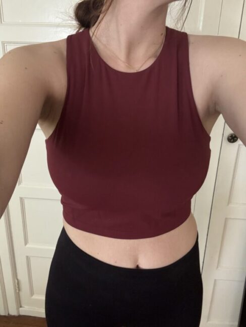 A person wearing a sleeveless maroon crop top and black pants standing indoors in front of a white door.