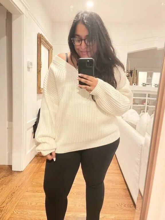 A woman with long dark hair and glasses takes a mirror selfie in a room, wearing a white oversized sweater and black leggings.