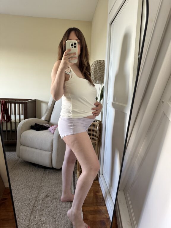 A woman in a white tank top and shorts takes a mirror selfie, holding her pregnant belly in a bedroom with a crib and armchair in the background.