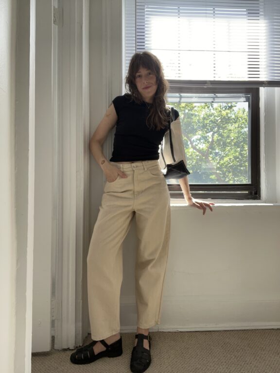A woman stands indoors in front of a window with blinds, wearing a black shirt, beige wide-leg pants, and black shoes. Sunlight streams through the window behind her.