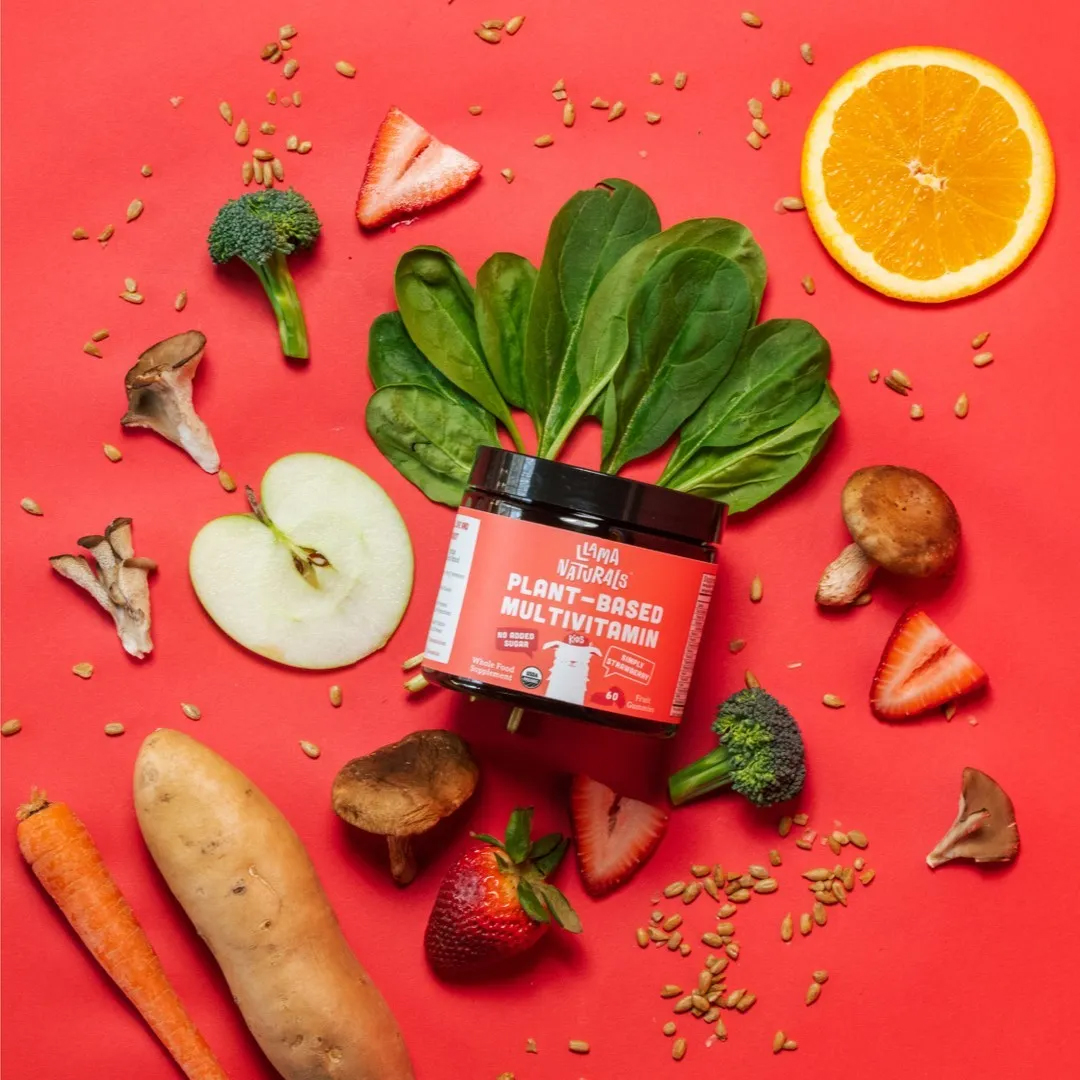 A styled flat lay shot of Llama Naturals multivitamin with cut fruits and veg on a red background