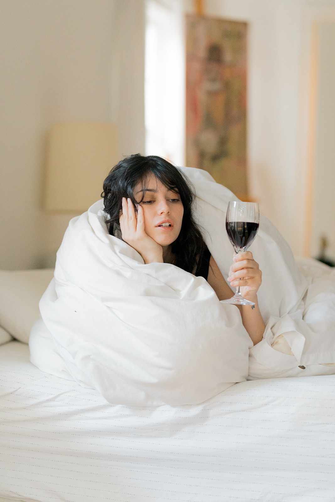 A woman wrapped in a white comforter sits on a bed, holding a glass of red wine and resting her head on her hand.