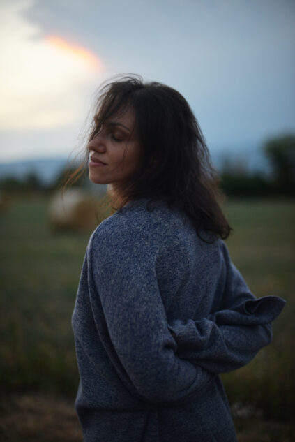 Woman in a gray sweater stands in a field at dusk with hay bales in the background, looking over her shoulder with eyes closed.