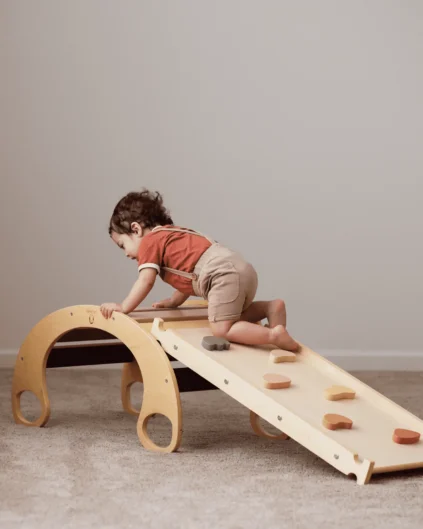 A toddler climbs a wooden play structure with a ramp and arch indoors, wearing a red shirt and suspenders.