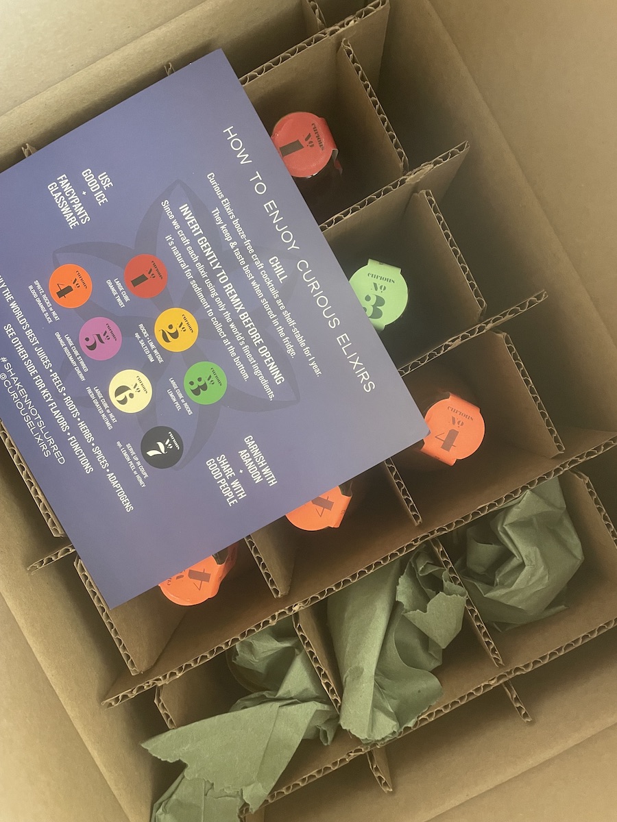An overhead image of an open box of Curious Elixirs bottled drinks, individually wrapped, with a description card on top of the package. 
