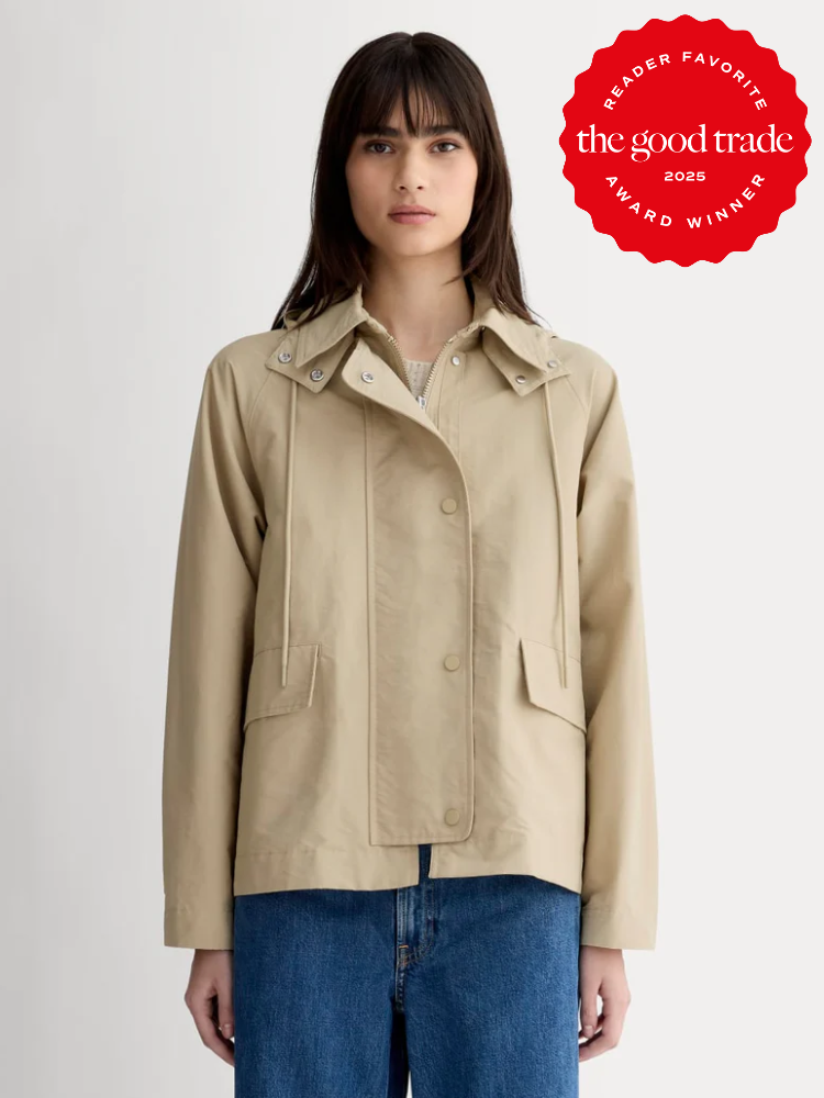 A woman wears a beige button-up jacket over a light top with blue jeans, standing against a plain background. A red "The Good Trade 2025 Award Winner" badge is in the top right corner.