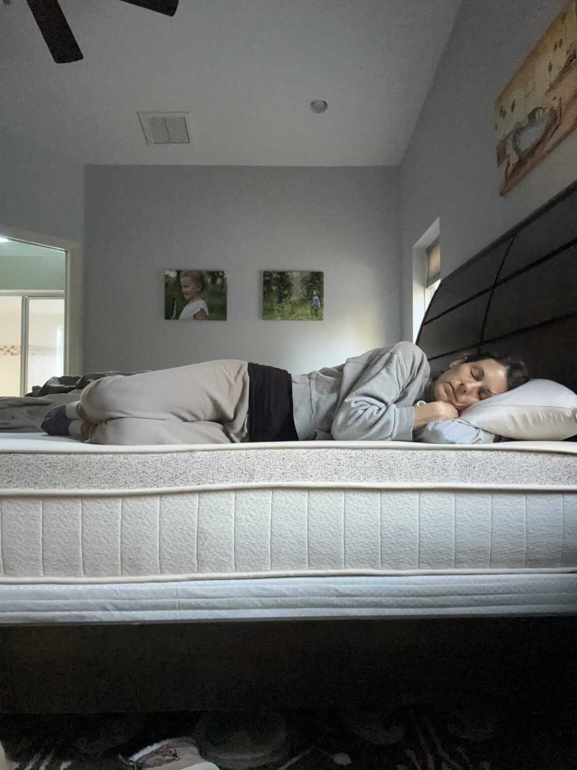 Person sleeping on their side in a bed with a white mattress and black frame in a softly lit room. Walls are light-colored with framed photos above.