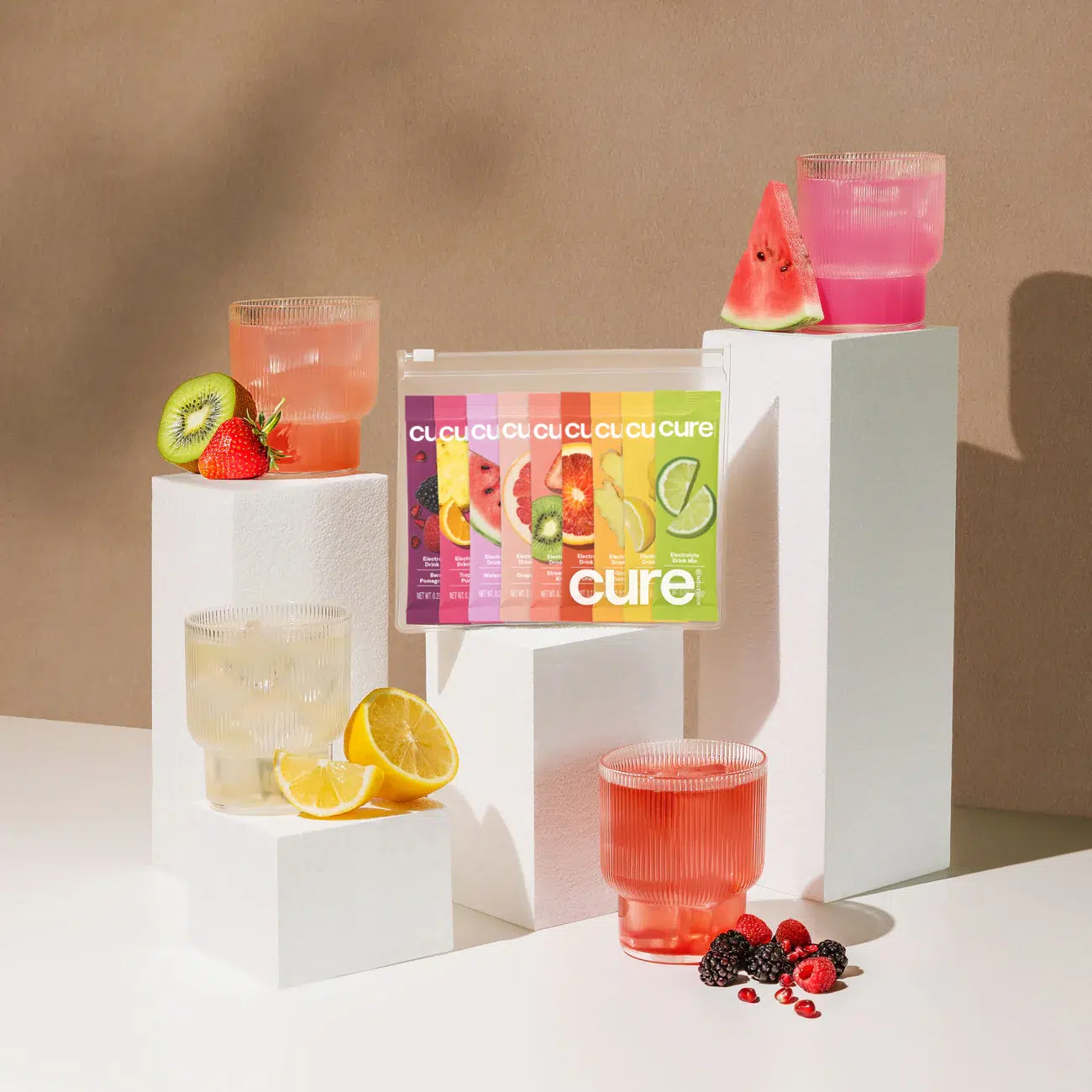 An arrangement of colorful drink packets and glasses filled with various fruit-infused beverages on geometric white blocks, surrounded by sliced kiwi, lemon, and berries.