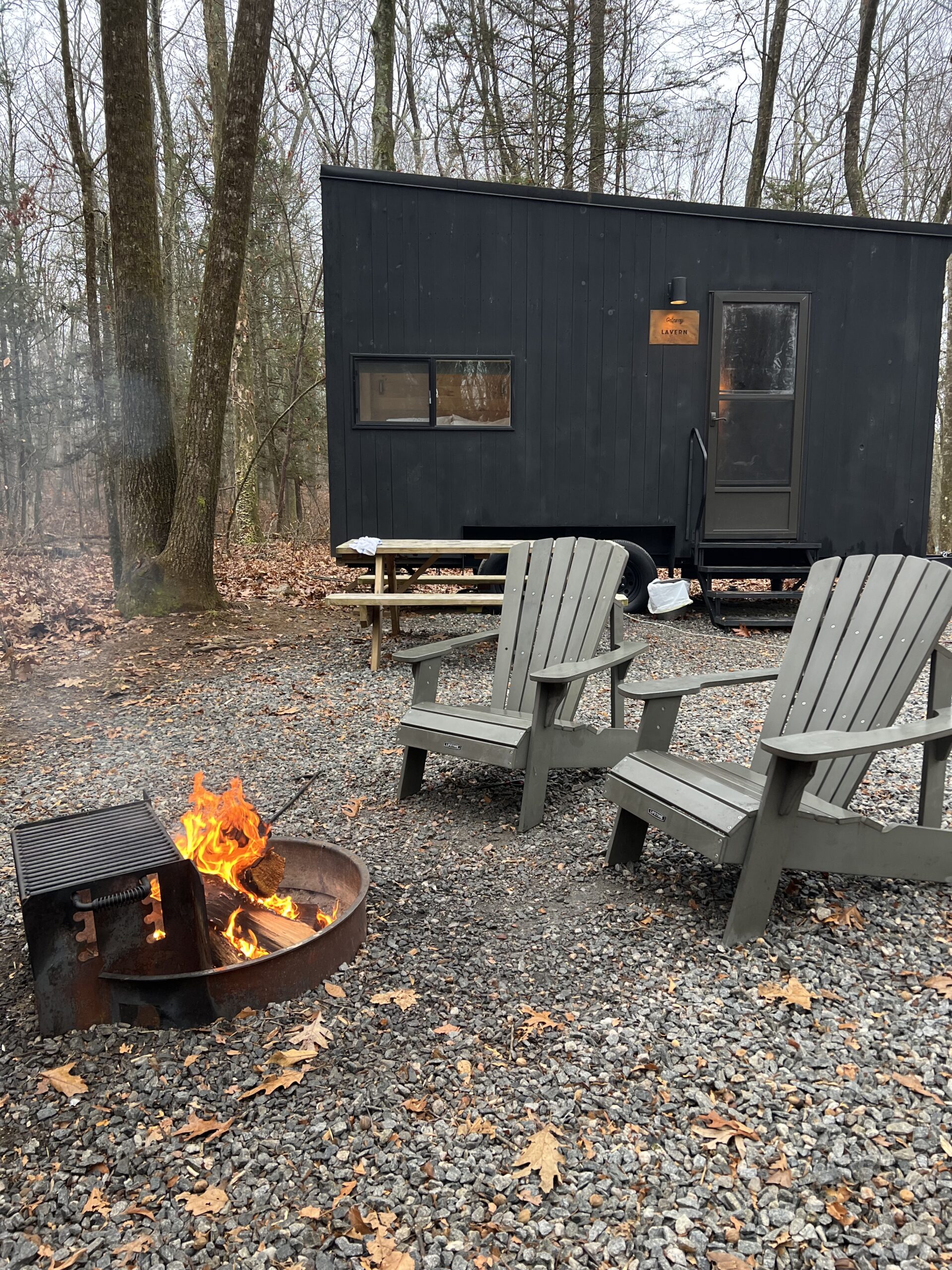 An exterior image of the black cabin with two Adirondack chairs and a firepit.