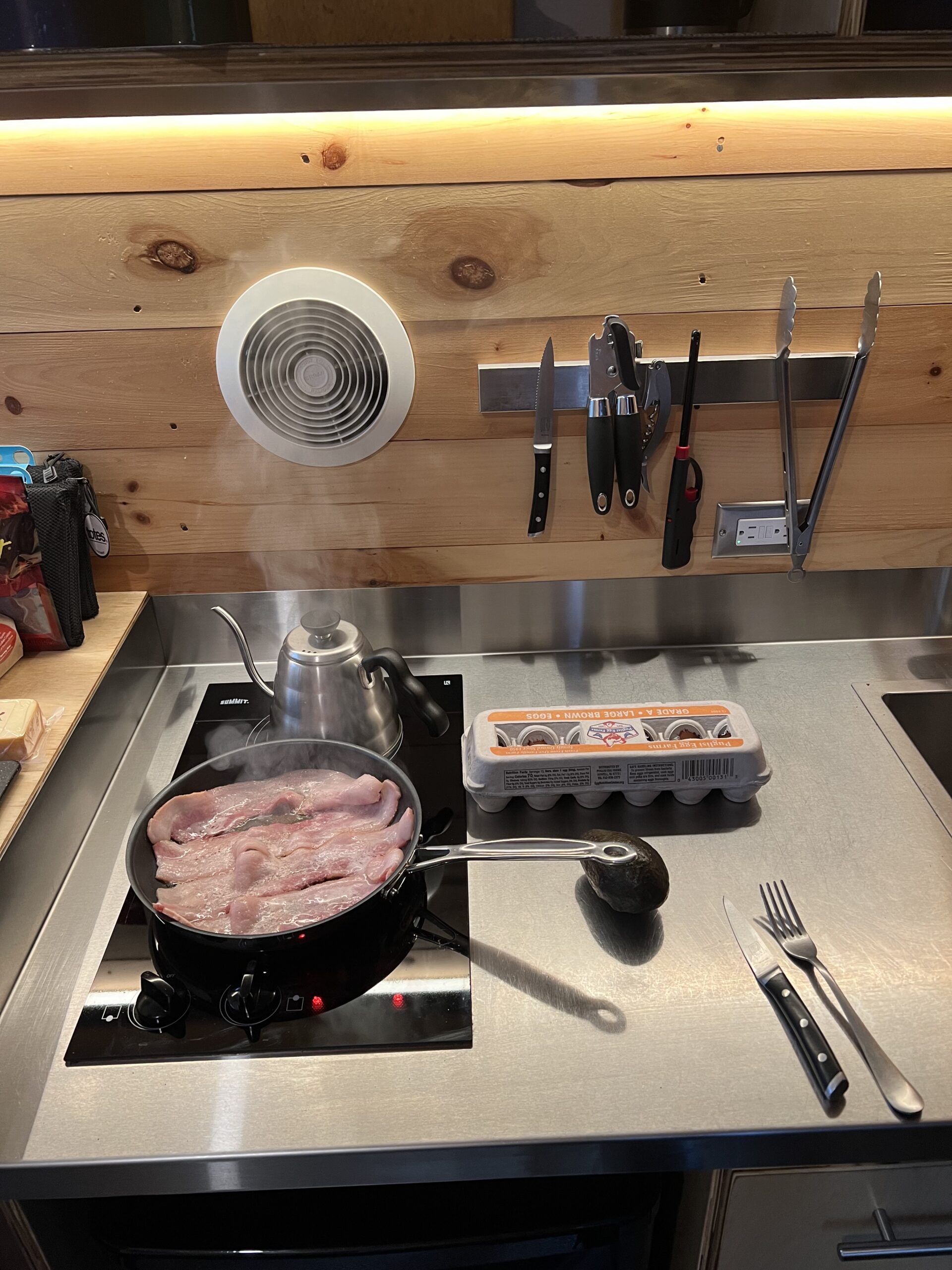 A stovetop with bacon frying in a pan.