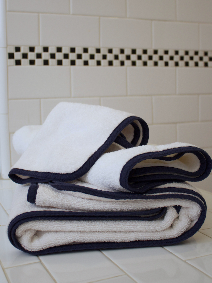 Two folded organic towels with dark trim are stacked on a white tiled surface in front of a tiled wall with a black and white checkered accent.