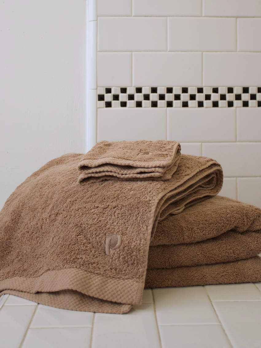 A neatly folded stack of organic towels and washcloths sits on a white tiled surface in front of a white tile wall with a black and white accent strip.