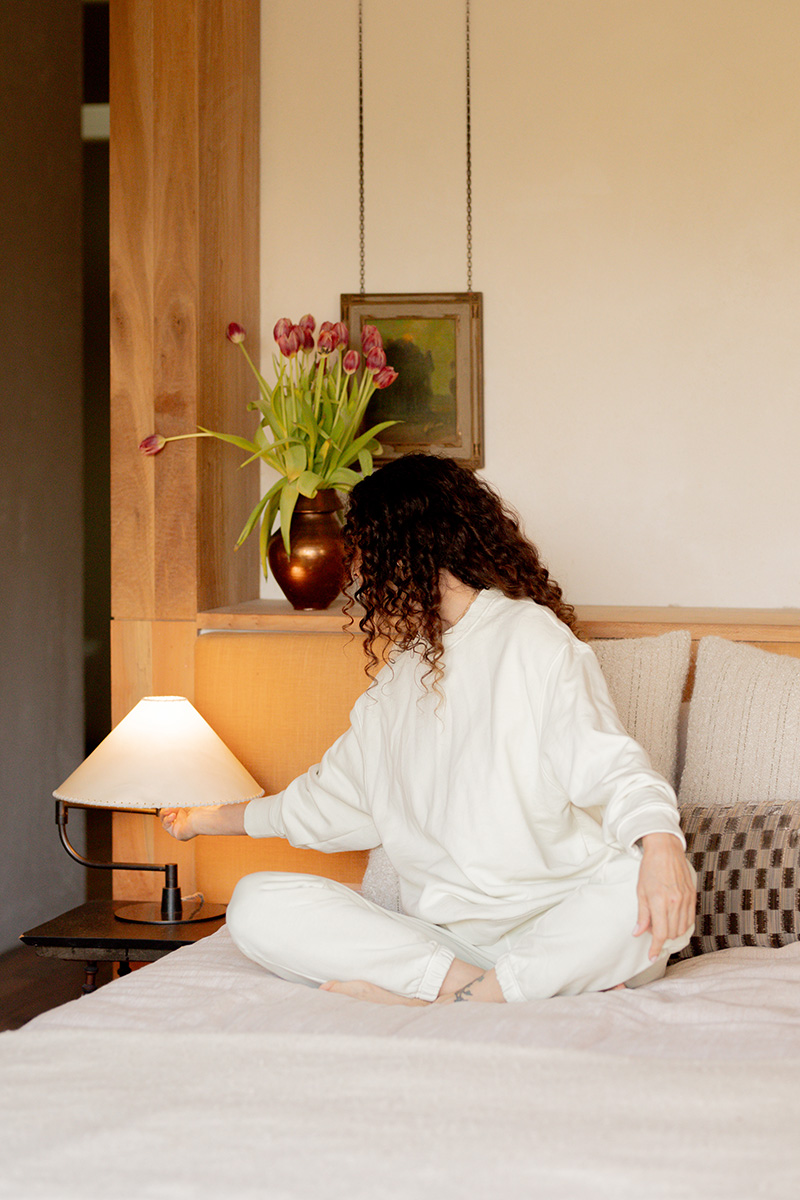 A woman in white loungewear sits cross-legged on a bed, facing away from the camera, reaching toward a table lamp beside a vase of tulips.