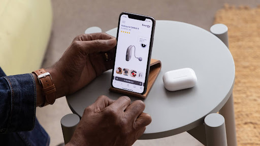 Person using a smartphone to shop for earphones online, with wireless earbuds and case on the table.